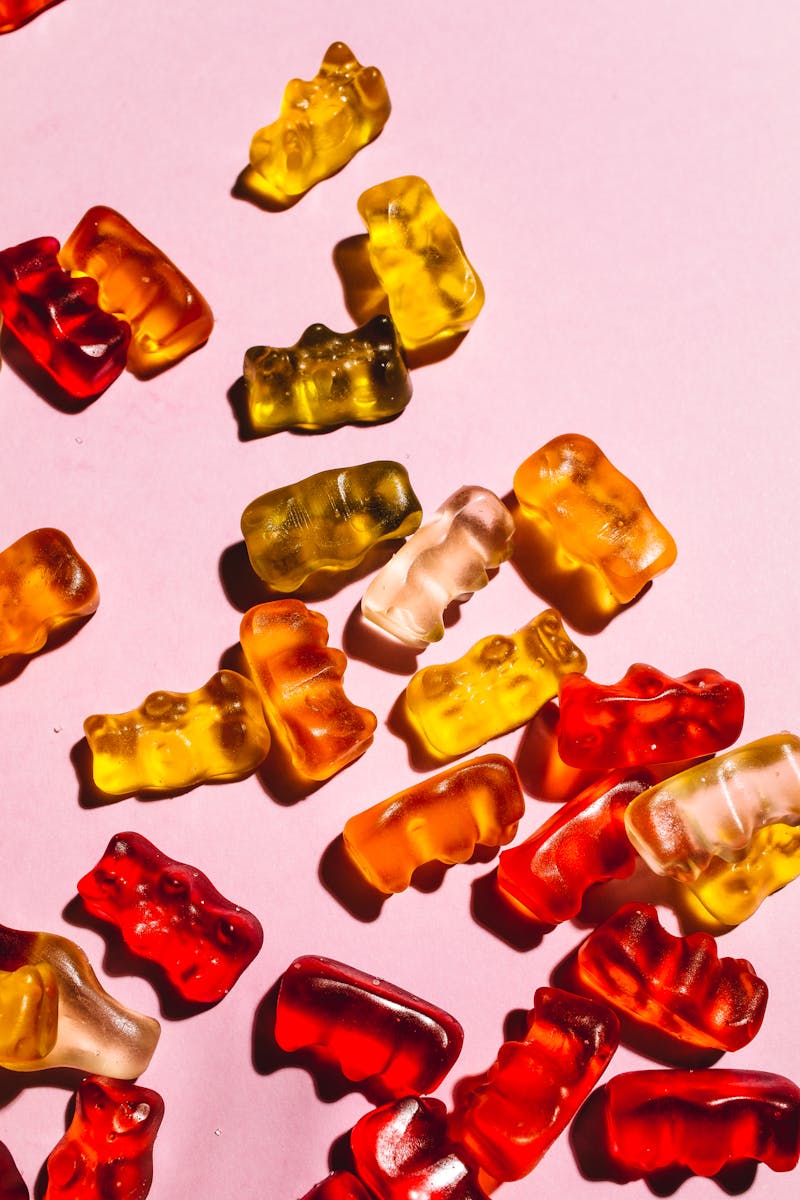 Colorful gummy bears spread across a pink background in bright lighting, creating a playful scene.