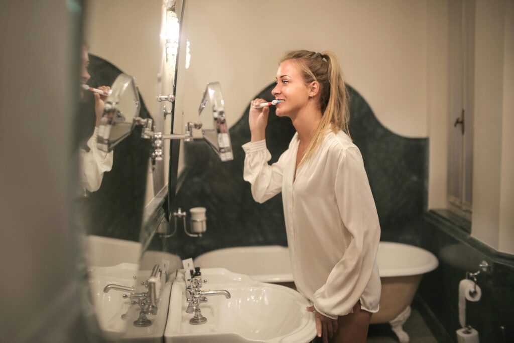 A cheerful young woman in a bathroom brushing her teeth, reflected in a mirror.