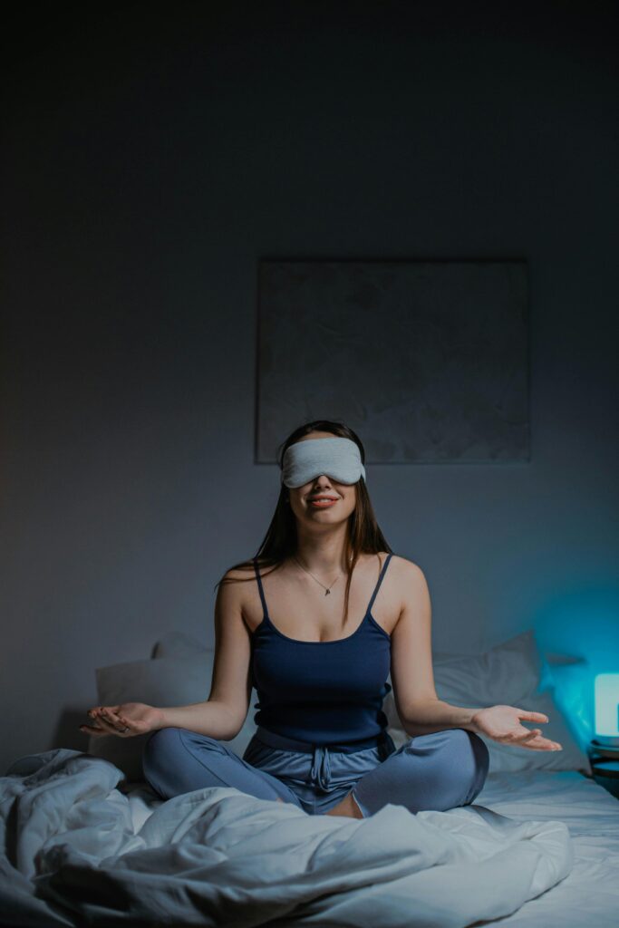 A woman sits cross-legged, meditating with a sleep mask on a bed, in a calm bedroom setting.