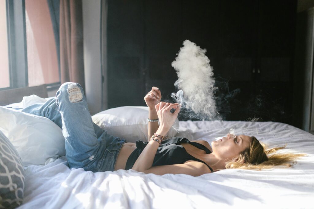 Woman lying on bed smoking in a casual bedroom environment, surrounded by natural light.